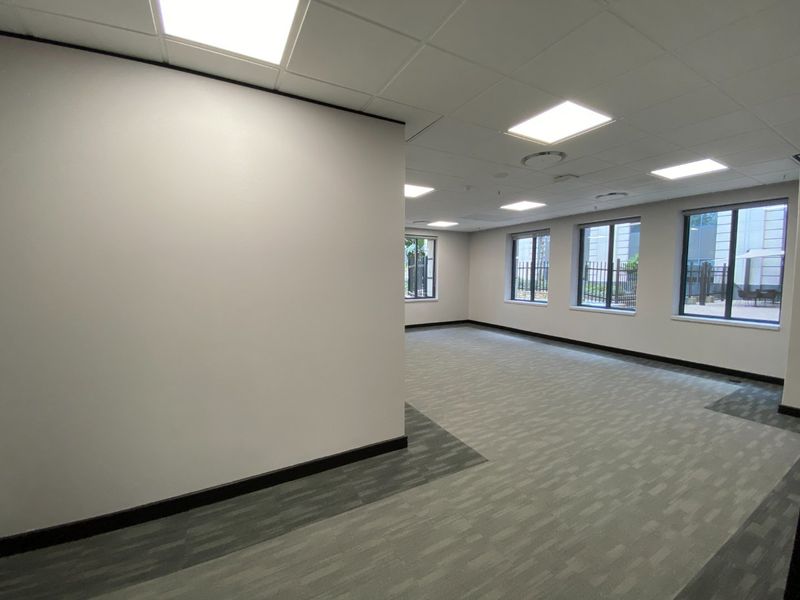 A bright, open-plan commercial office space featuring neutral carpet tiles, recessed ceiling lighting, and large windows providing ample natural light.