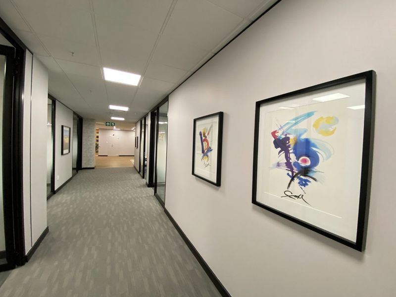A modern, well-maintained commercial office hallway featuring glass partitions, recessed ceiling lighting, and neutral carpet flooring.
