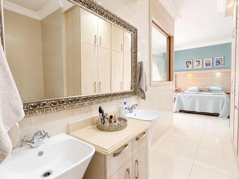 Modern open-plan bathroom featuring dual vanity basins, extensive built-in cabinetry, and large wall mirror, seamlessly flowing into the adjacent bedroom area.