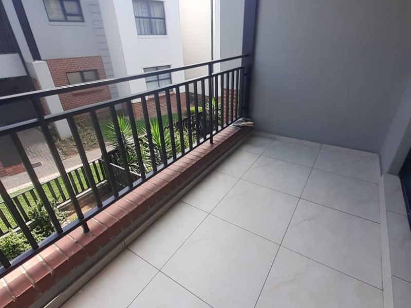 A clean, tiled balcony featuring a metal balustrade and a brick ledge, overlooking the apartment complex grounds.