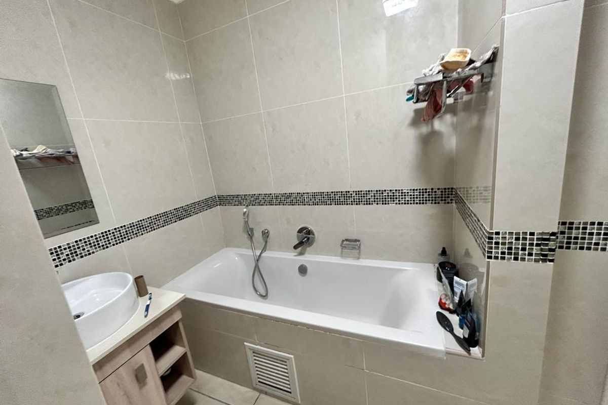 A functional bathroom featuring a built-in bathtub with handheld shower mixer, a vanity cabinet with a vessel sink, and neutral wall tiling with mosaic accents.