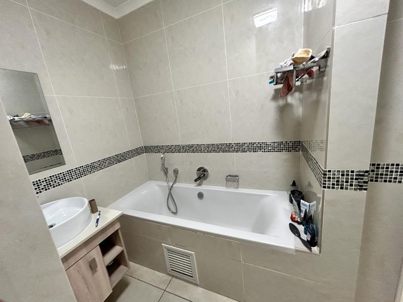 A functional bathroom featuring a built-in bathtub with handheld shower mixer, a vanity cabinet with a vessel sink, and neutral wall tiling with mosaic accents.