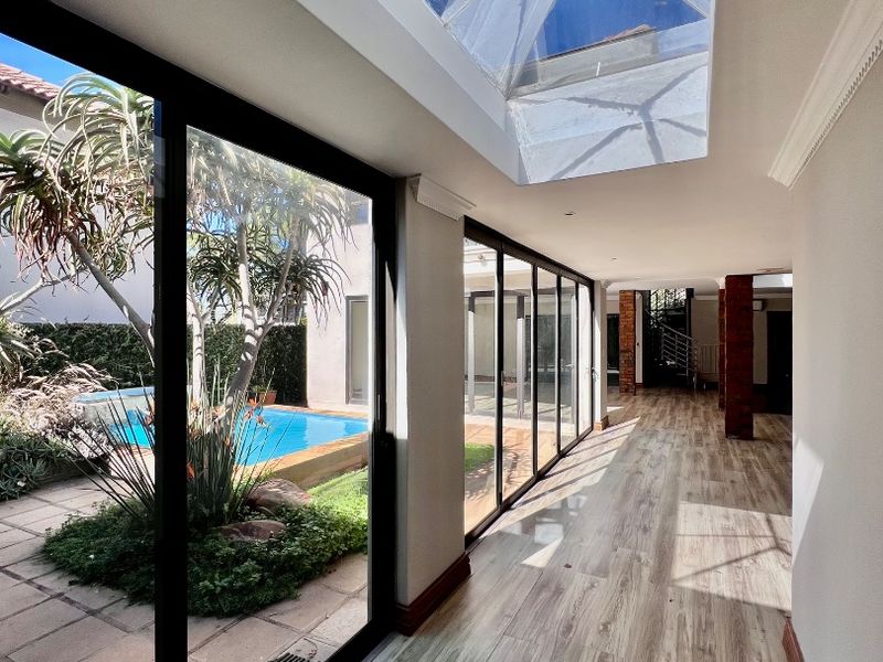A bright, modern hallway featuring extensive glass sliding doors, a large skylight, and wood-look laminate flooring, offering a seamless view of the outdoor pool area.