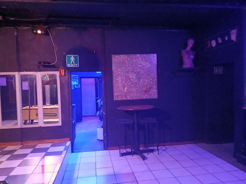 A dimly lit bar area featuring dark walls, tiled flooring, and an exit point leading to an adjacent space.