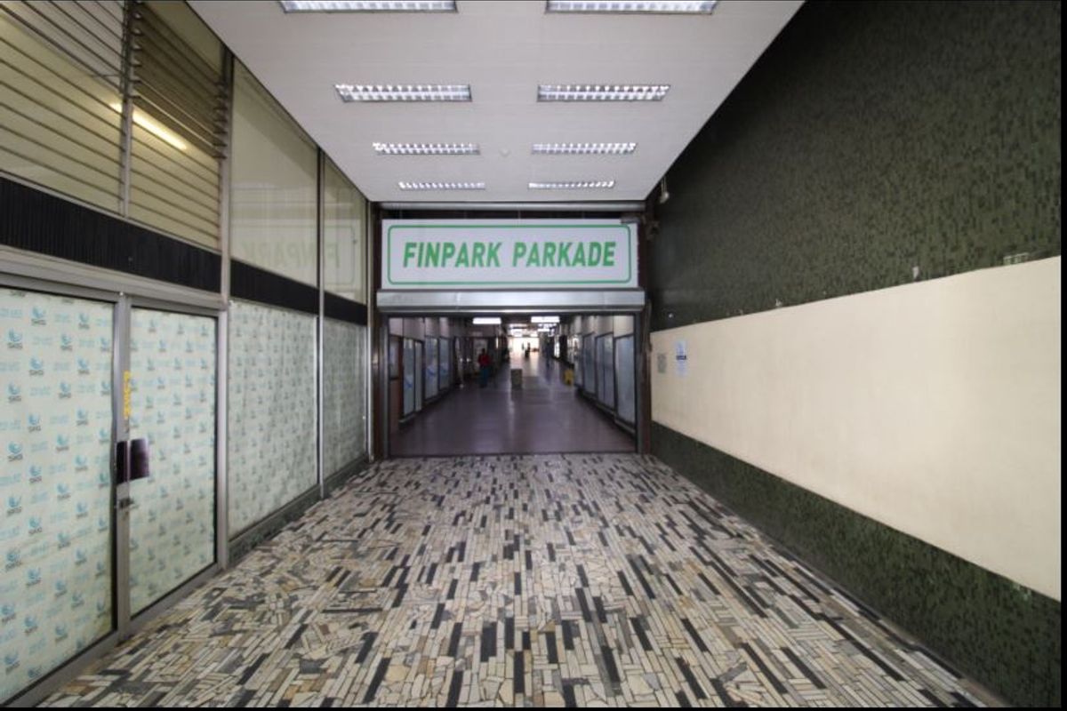 A commercial hallway or pedestrian walkway featuring tiled flooring, fluorescent lighting, and glass storefront access points leading toward the parking facility.