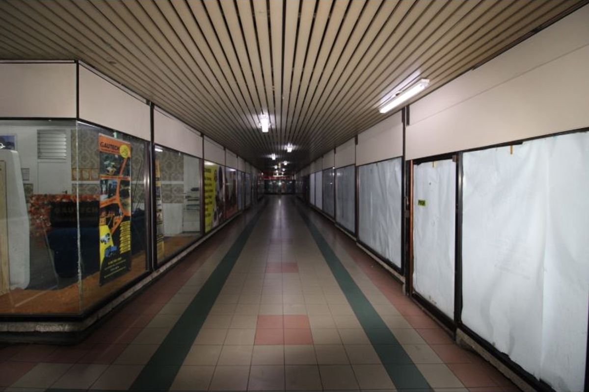 A commercial hallway or corridor featuring a slatted ceiling, fluorescent lighting, and multiple glass-fronted retail units along the walkway.
