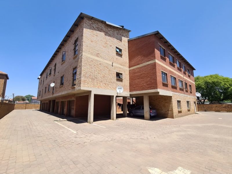 Exterior view of a multi-story face brick apartment building featuring covered ground-level parking bays and a paved driveway area.