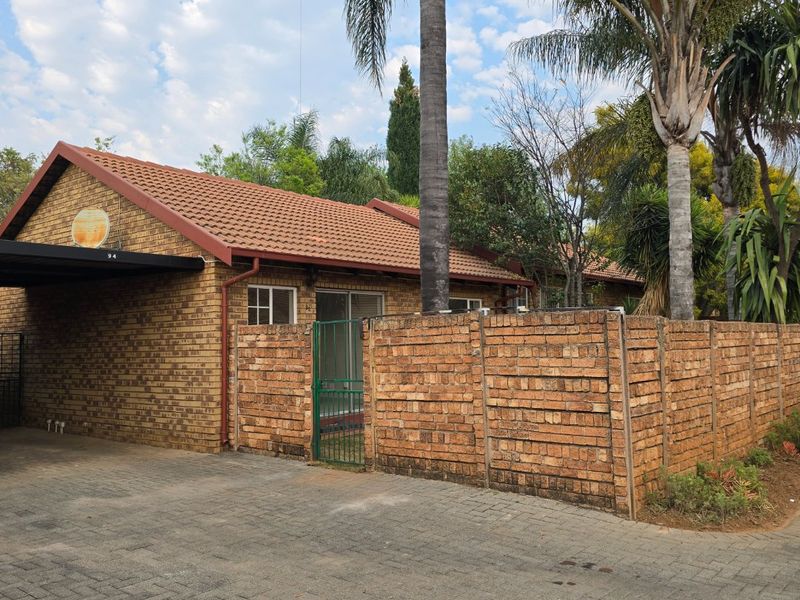 Front view of a facebrick townhouse featuring a tiled roof, paved driveway, integrated carport, and a secure gated entrance with boundary walling.