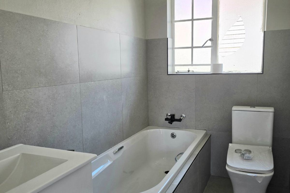A modern bathroom featuring a built-in bathtub, toilet, and vanity basin with neutral grey tiling throughout.