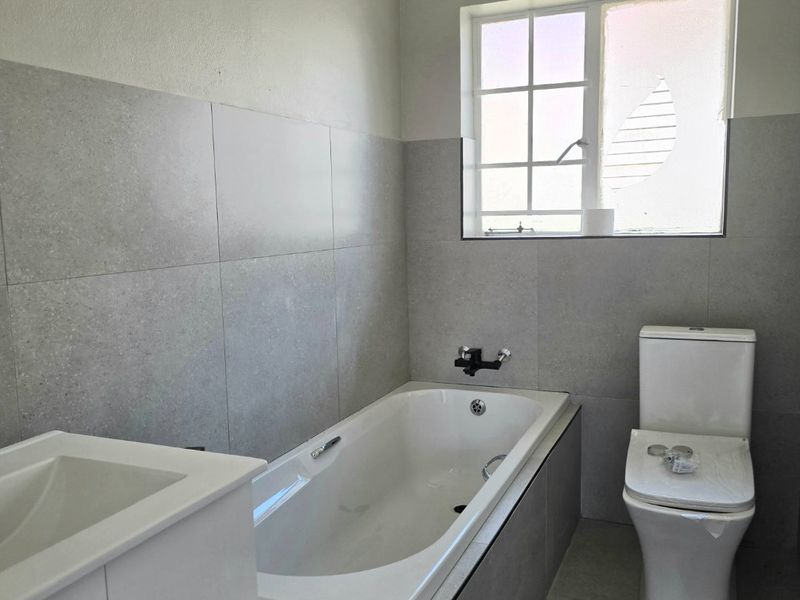 A modern bathroom featuring a built-in bathtub, toilet, and vanity basin with neutral grey tiling throughout.