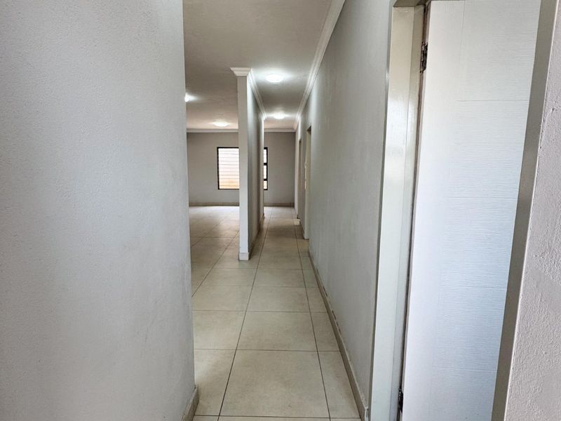 A bright, empty hallway featuring neutral tiled flooring, recessed lighting, and clean white walls leading into an open-plan living area.