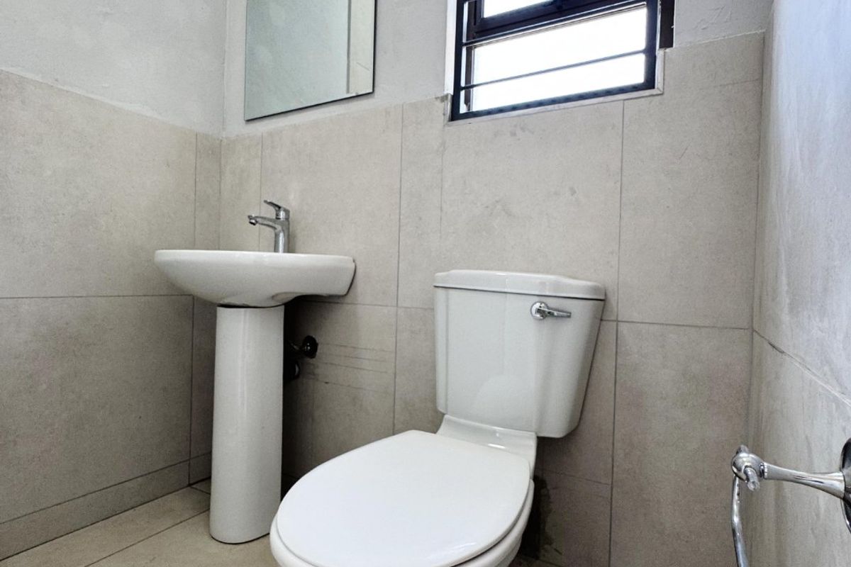 A functional bathroom featuring a pedestal basin, toilet, and neutral wall and floor tiling with a window for natural light.