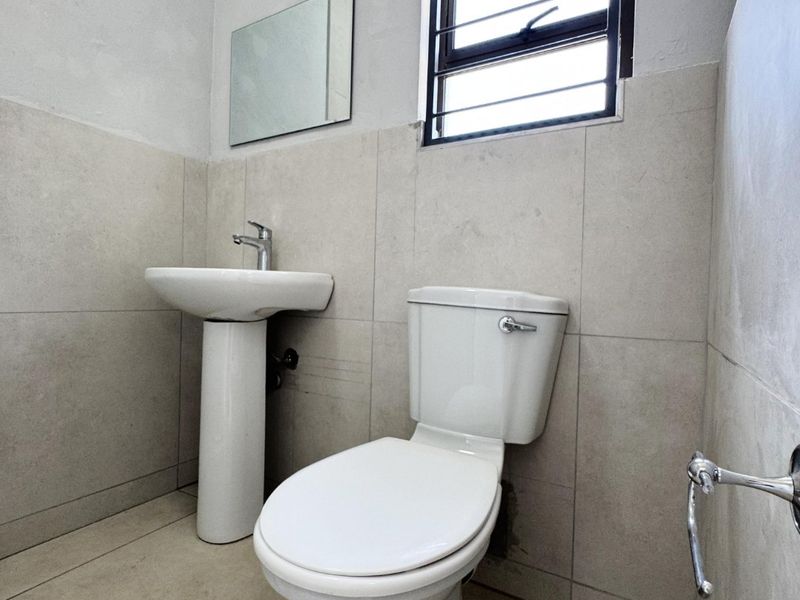 A functional bathroom featuring a pedestal basin, toilet, and neutral wall and floor tiling with a window for natural light.