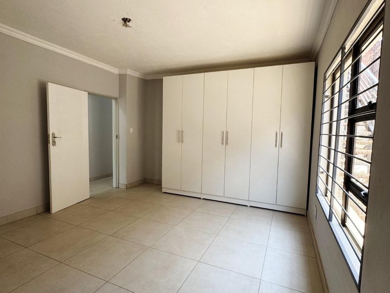 A bright, empty bedroom featuring neutral tiled flooring, built-in white cupboards, and a window fitted with security bars.