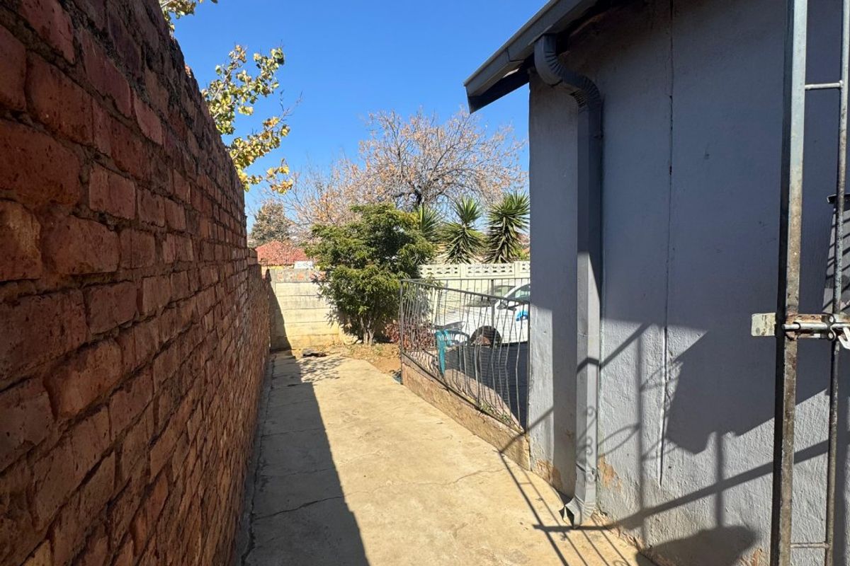 A narrow side passage featuring a concrete walkway, brick boundary wall, and exterior building wall with a metal security gate.