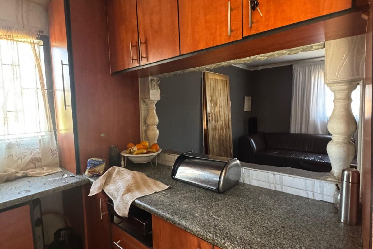 A dated kitchen featuring laminate cabinetry, granite countertops, and decorative pillars, with a pass-through window looking into the adjacent living area.