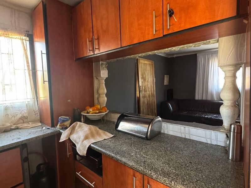 A dated kitchen featuring laminate cabinetry, granite countertops, and decorative pillars, with a pass-through window looking into the adjacent living area.