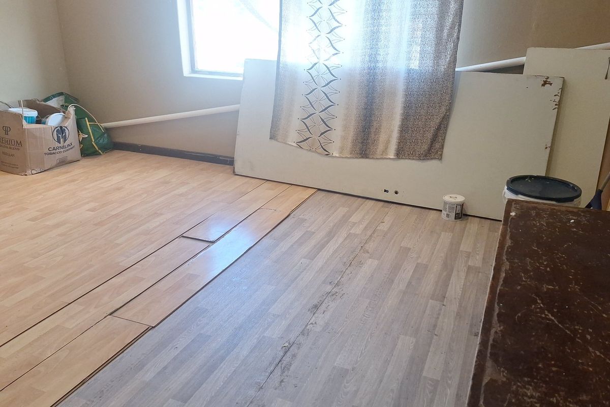 An unfinished bedroom space featuring mismatched laminate flooring, exposed piping, and visible wall repairs, requiring completion and cosmetic updates.