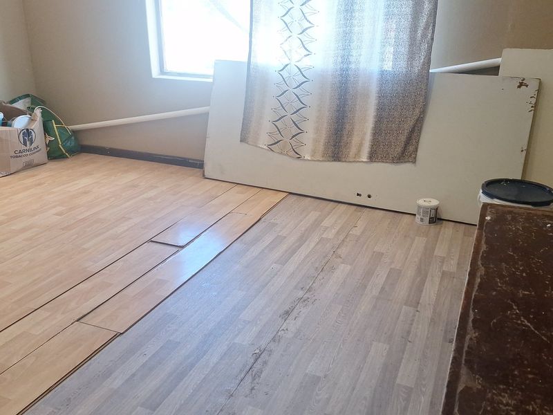 An unfinished bedroom space featuring mismatched laminate flooring, exposed piping, and visible wall repairs, requiring completion and cosmetic updates.