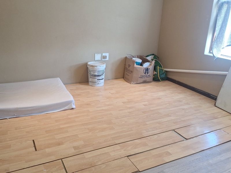 A bedroom space featuring light-toned laminate flooring with visible installation gaps and an exposed wall pipe, requiring finishing and maintenance.