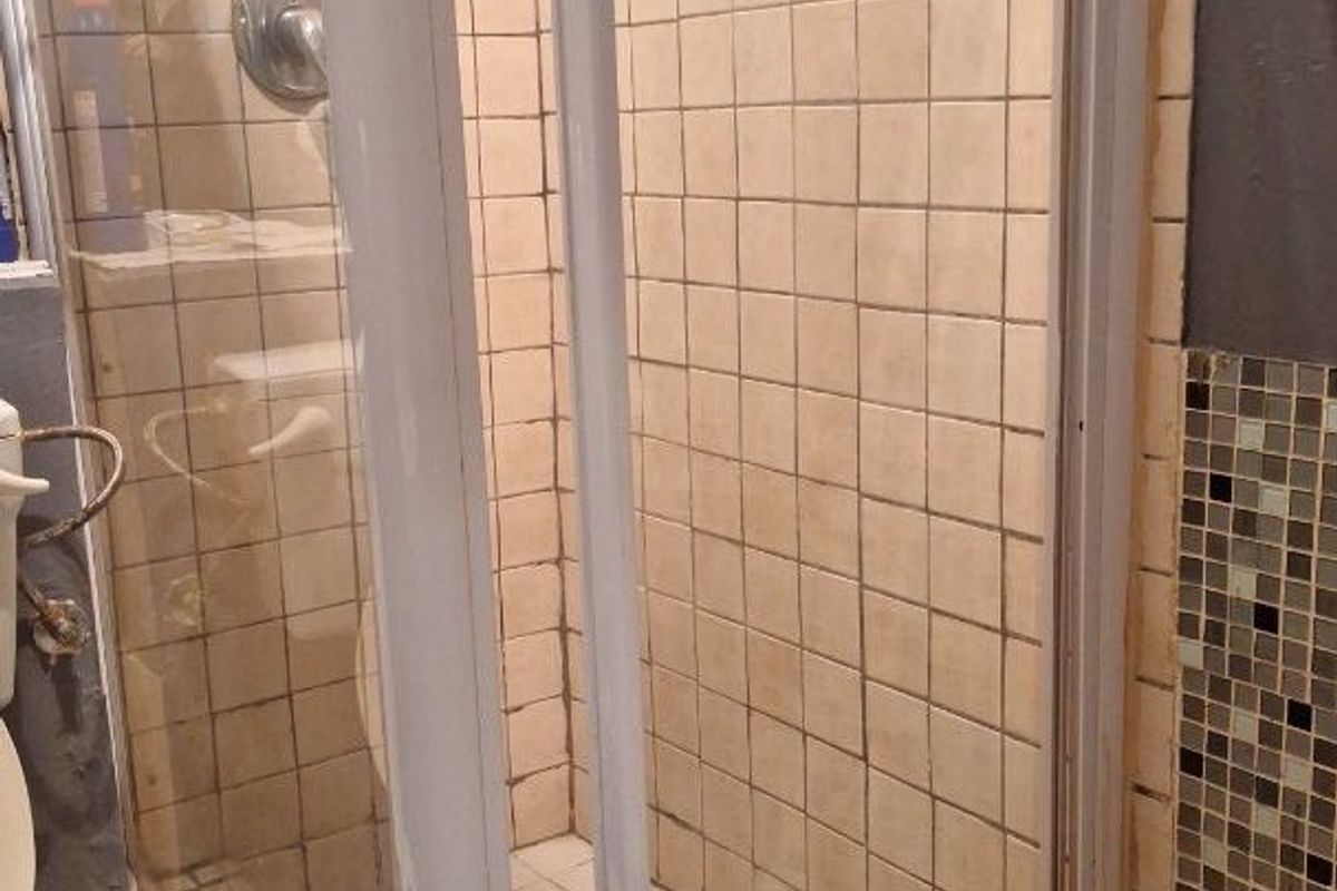 A bathroom featuring a shower enclosure with tiled walls and flooring, alongside a toilet. The shower door frame shows signs of damage.