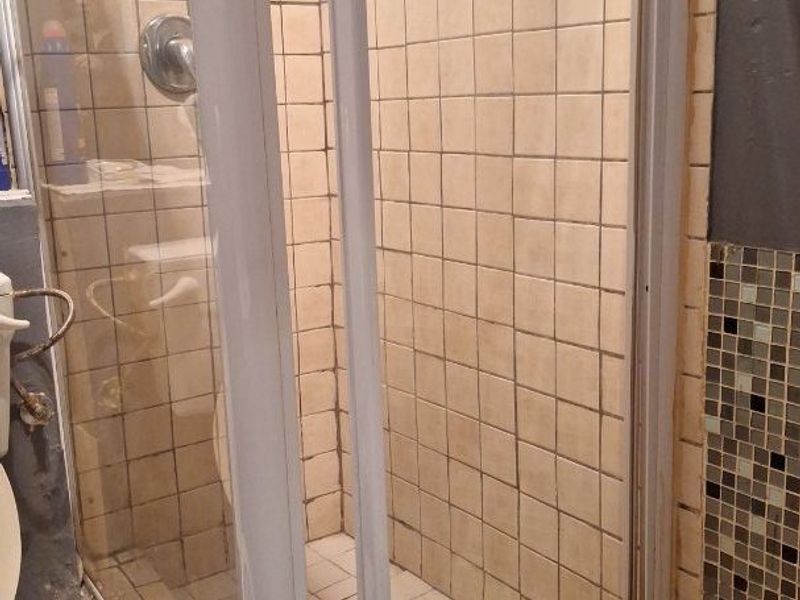 A bathroom featuring a shower enclosure with tiled walls and flooring, alongside a toilet. The shower door frame shows signs of damage.