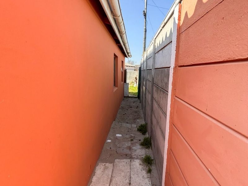 A narrow side passage between the house exterior and a precast concrete boundary wall, featuring basic paving and guttering.