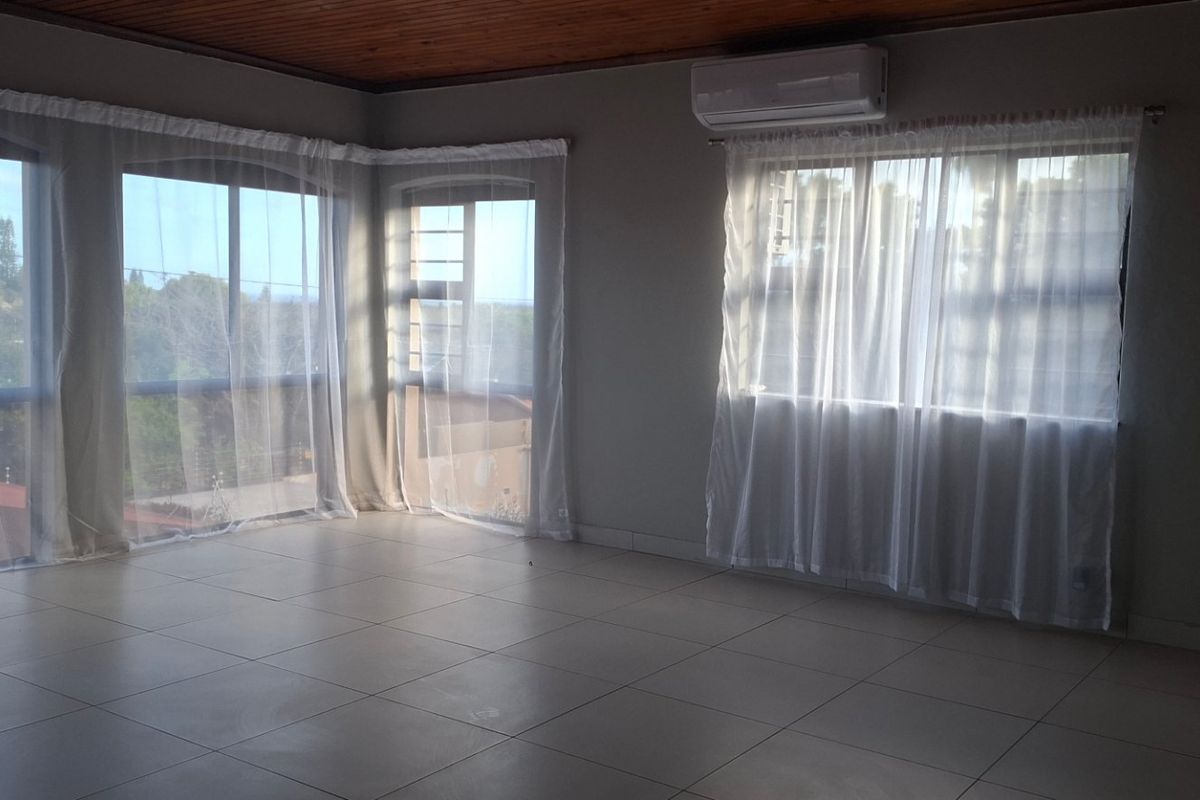 A spacious, empty living area featuring tiled flooring, a timber-paneled ceiling, and a wall-mounted air conditioning unit with large windows providing natural light.