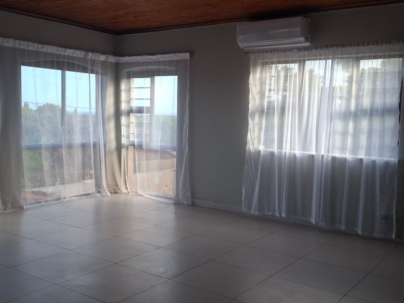 A spacious, empty living area featuring tiled flooring, a timber-paneled ceiling, and a wall-mounted air conditioning unit with large windows providing natural light.