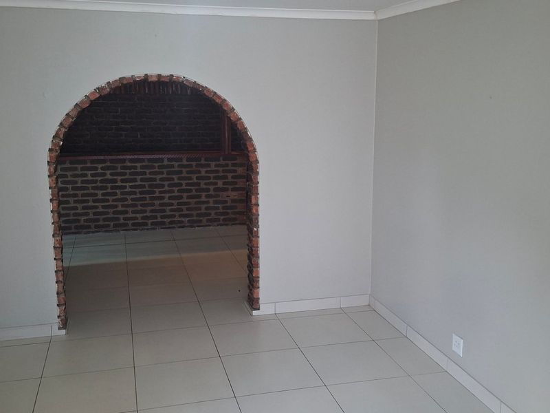 An empty living area featuring neutral wall tones, tiled flooring, and a distinctive exposed brick archway leading to an adjacent space.