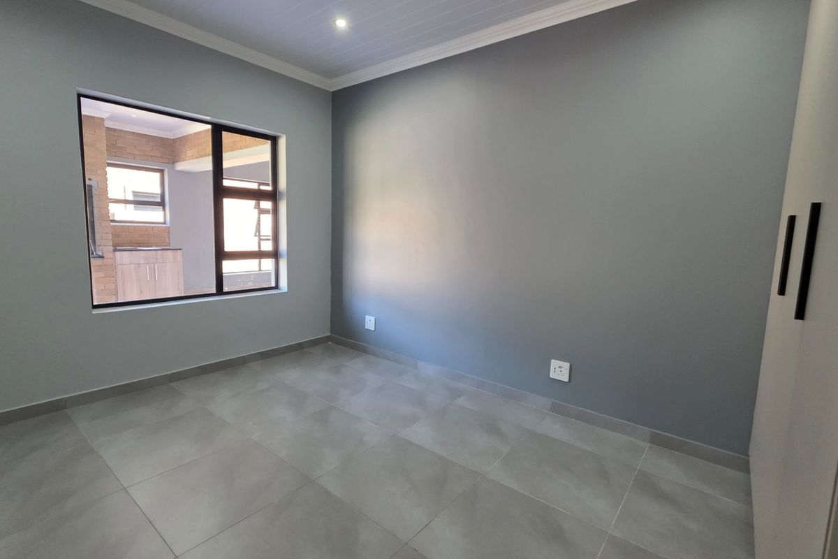 A modern, empty bedroom featuring neutral grey walls, large-format floor tiles, built-in cupboards, and a large window providing natural light.