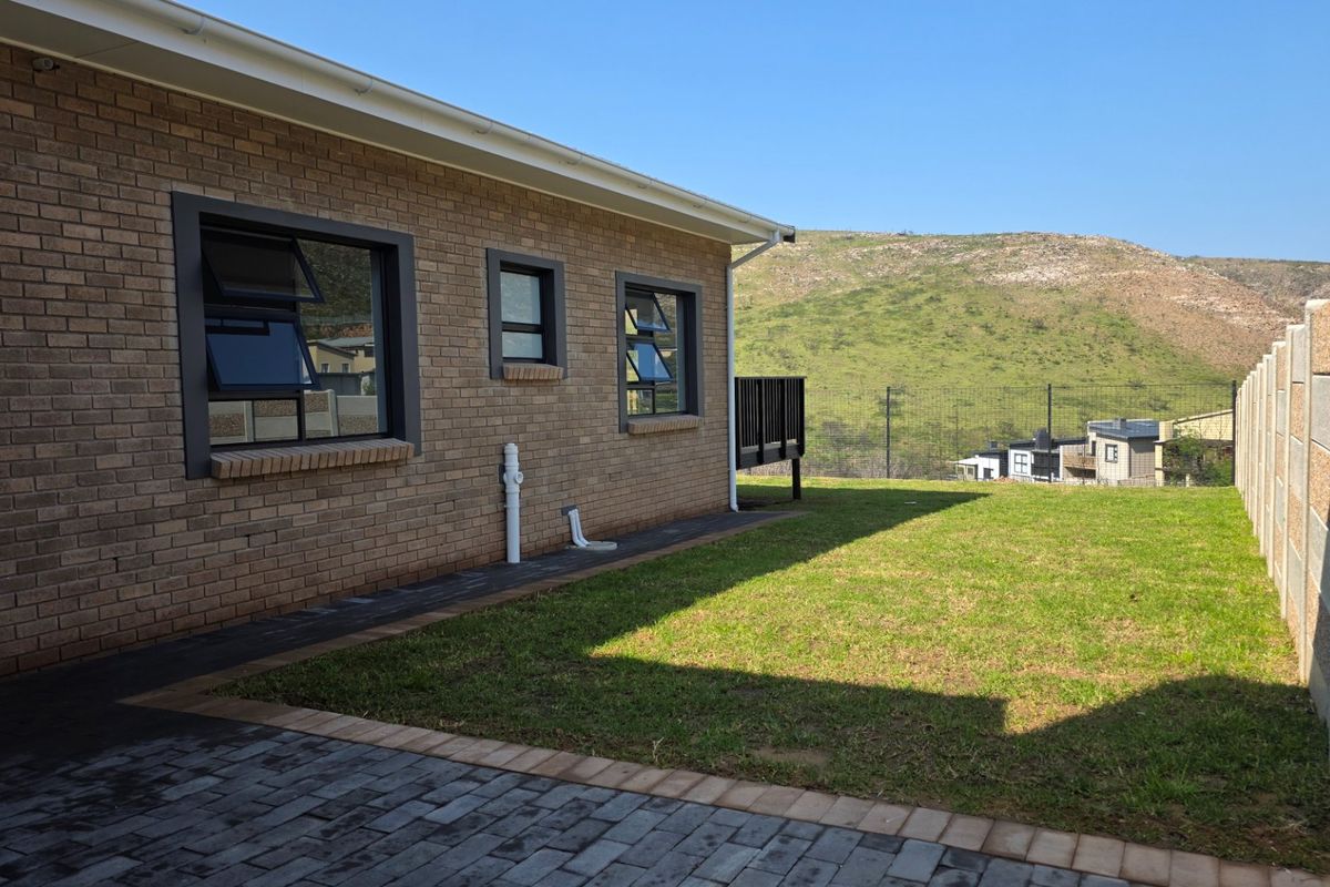 Rear exterior view of the house featuring a brick facade, paved walkway, well-maintained lawn, and a precast concrete boundary wall with scenic hill views.