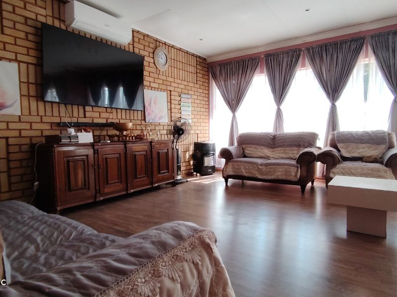 A living room featuring an exposed brick feature wall, laminate flooring, and a wall-mounted air conditioning unit with large windows allowing natural light.