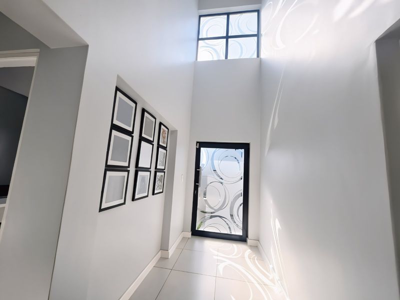 A bright, modern double-volume entrance hall featuring high ceilings, a large clerestory window, and a contemporary frosted glass front door with tiled flooring.