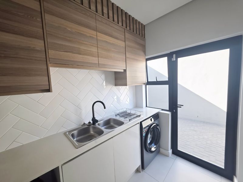 Modern laundry room featuring sleek wood-look cabinetry, a herringbone tiled backsplash, double stainless steel sink, and direct access to an outdoor area.