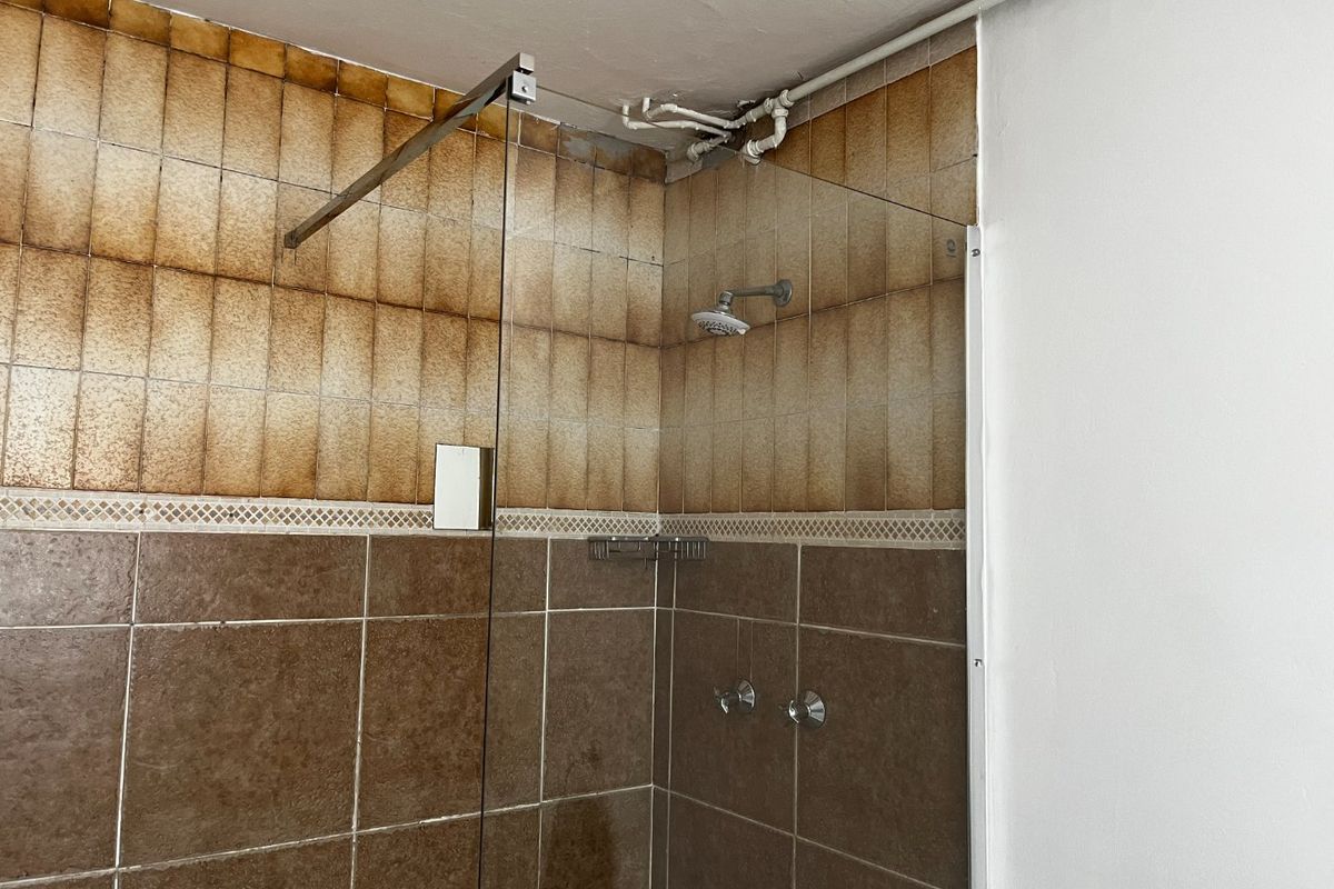 A shower enclosure featuring a glass screen and tiled walls with visible staining and exposed surface-mounted plumbing.