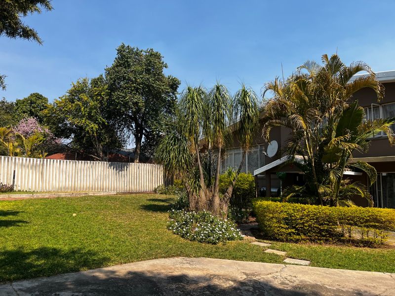 A rear garden view featuring a well-maintained lawn, mature landscaping, concrete boundary wall with electric fencing, and a paved patio area.
