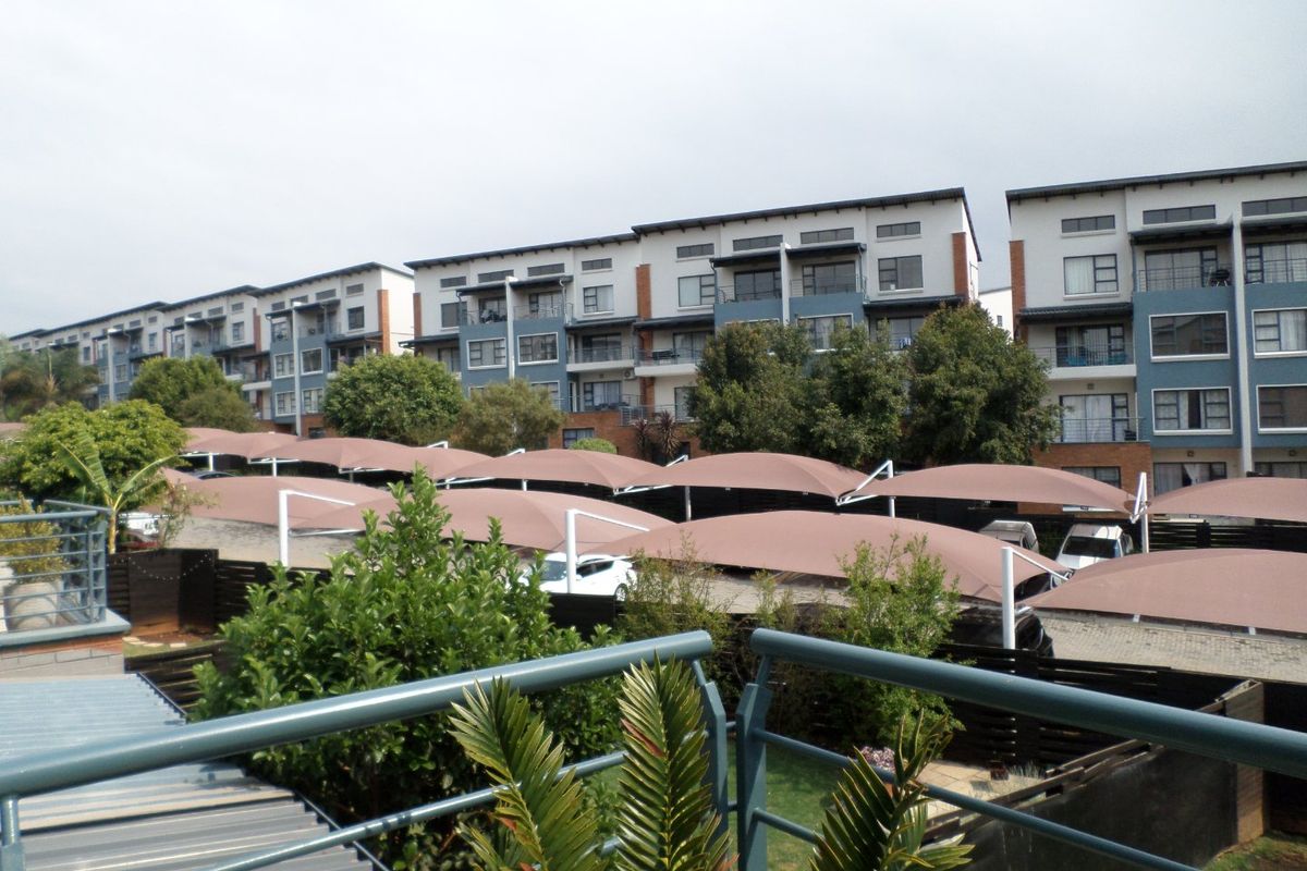 View of the apartment complex exterior featuring covered parking bays, landscaped garden areas, and a metal balcony railing in the foreground.