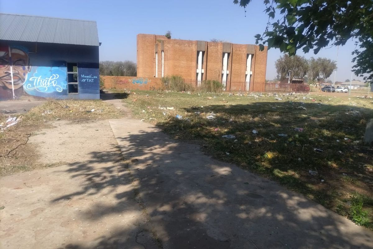 View of a vacant residential plot featuring a concrete path, brick boundary wall, and a nearby brick building with visible graffiti and debris.