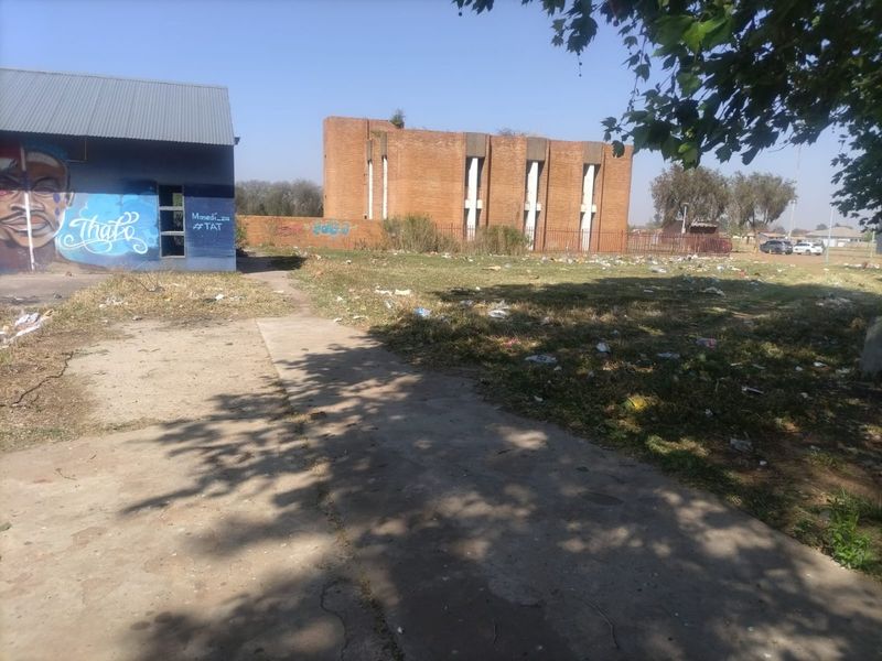 View of a vacant residential plot featuring a concrete path, brick boundary wall, and a nearby brick building with visible graffiti and debris.
