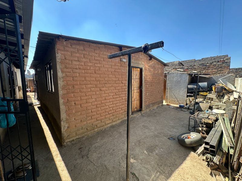 Rear exterior view of a brick structure featuring a metal security door, corrugated roof, and concrete paved yard area.