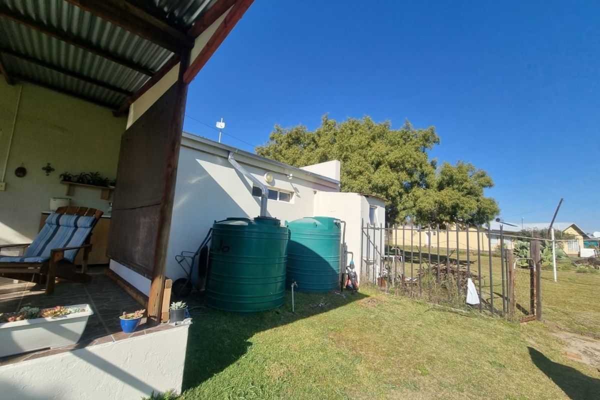 Rear exterior view showing a covered patio area, water storage tanks, and metal security fencing set against a garden backdrop.