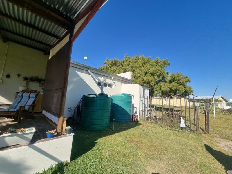 Rear exterior view showing a covered patio area, water storage tanks, and metal security fencing set against a garden backdrop.