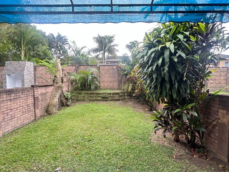 A private rear garden featuring a lawn, brick boundary walls, and a built-in braai area under a blue shade cloth awning.