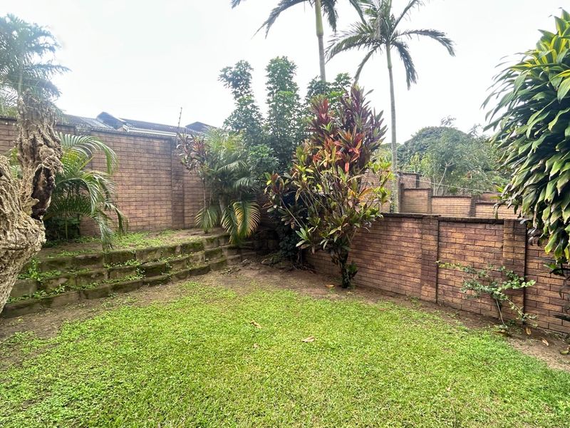 A private rear garden featuring established trees, a lawn area, and brick boundary walls with a tiered retaining wall structure.