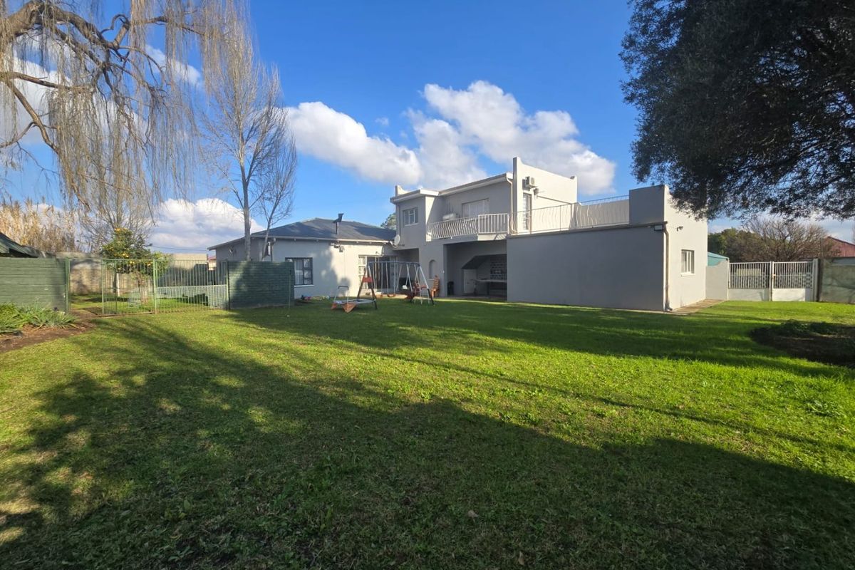 Rear view of the property featuring a spacious lawn, a two-story building section with an upper balcony, and a built-in outdoor braai area.