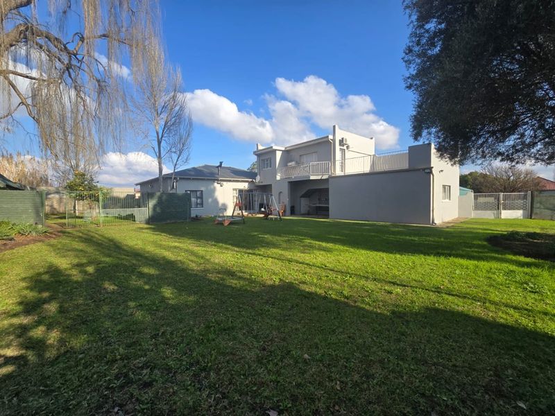 Rear view of the property featuring a spacious lawn, a two-story building section with an upper balcony, and a built-in outdoor braai area.