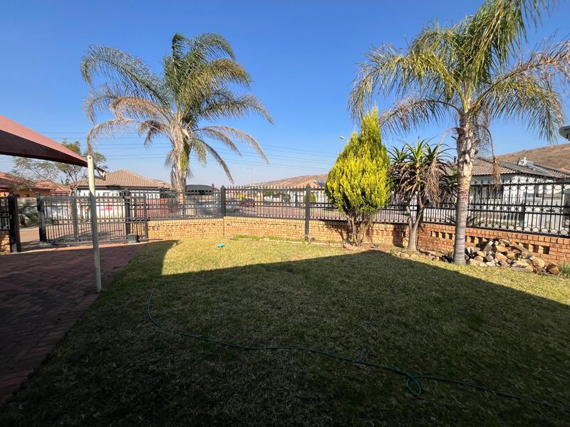 A well-maintained garden area featuring a brick boundary wall, wrought iron fencing, paved patio space, and established palm trees.