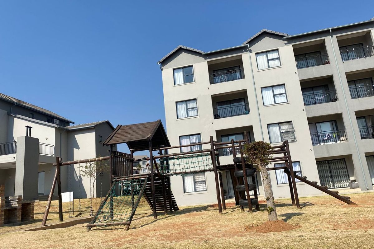 Exterior view of an apartment complex featuring a communal playground area with wooden climbing structures and a brick braai facility.