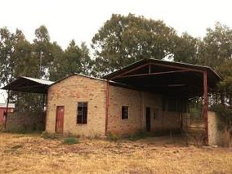 A rustic farm outbuilding featuring brick construction with extended metal roof coverings and steel support structures, set in a rural landscape.